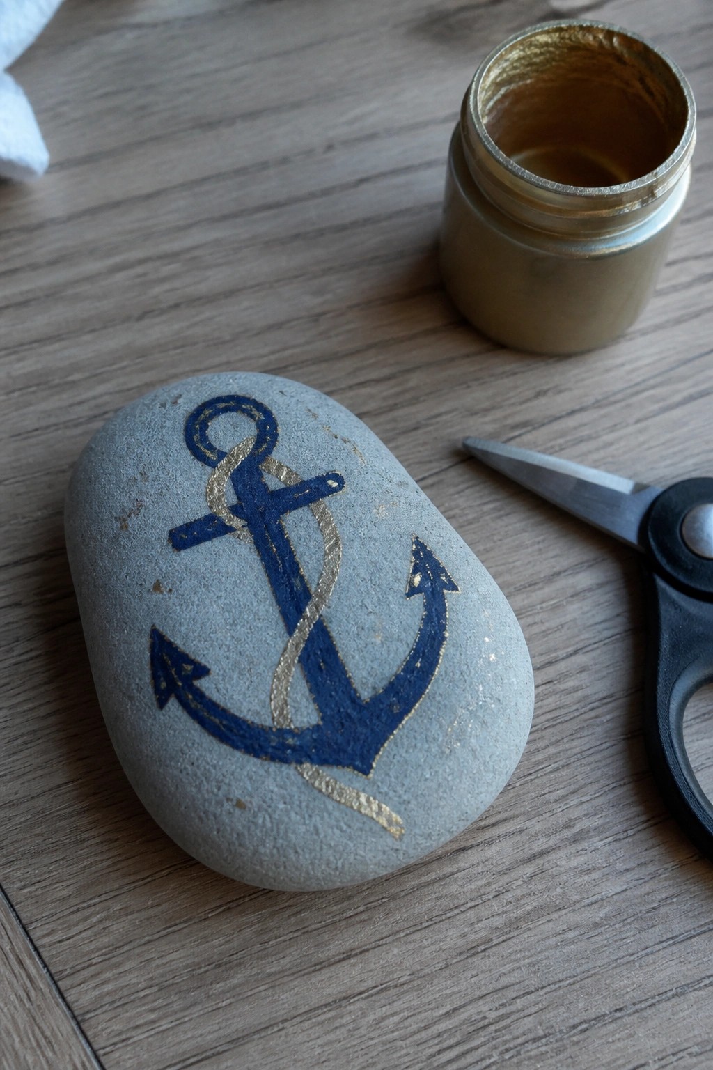 Gray stone with blue anchor and gold accents beside gold paint jar and scissors
