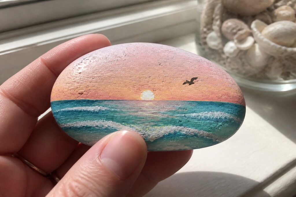 23 Brilliant Rock Painting Ideas for Beach Lovers Using Smooth Coastal Stones A hand holds a smooth oval rock painted with a sunset sky over teal waves, a small white sun on the horizon, and one black bird silhouette.
