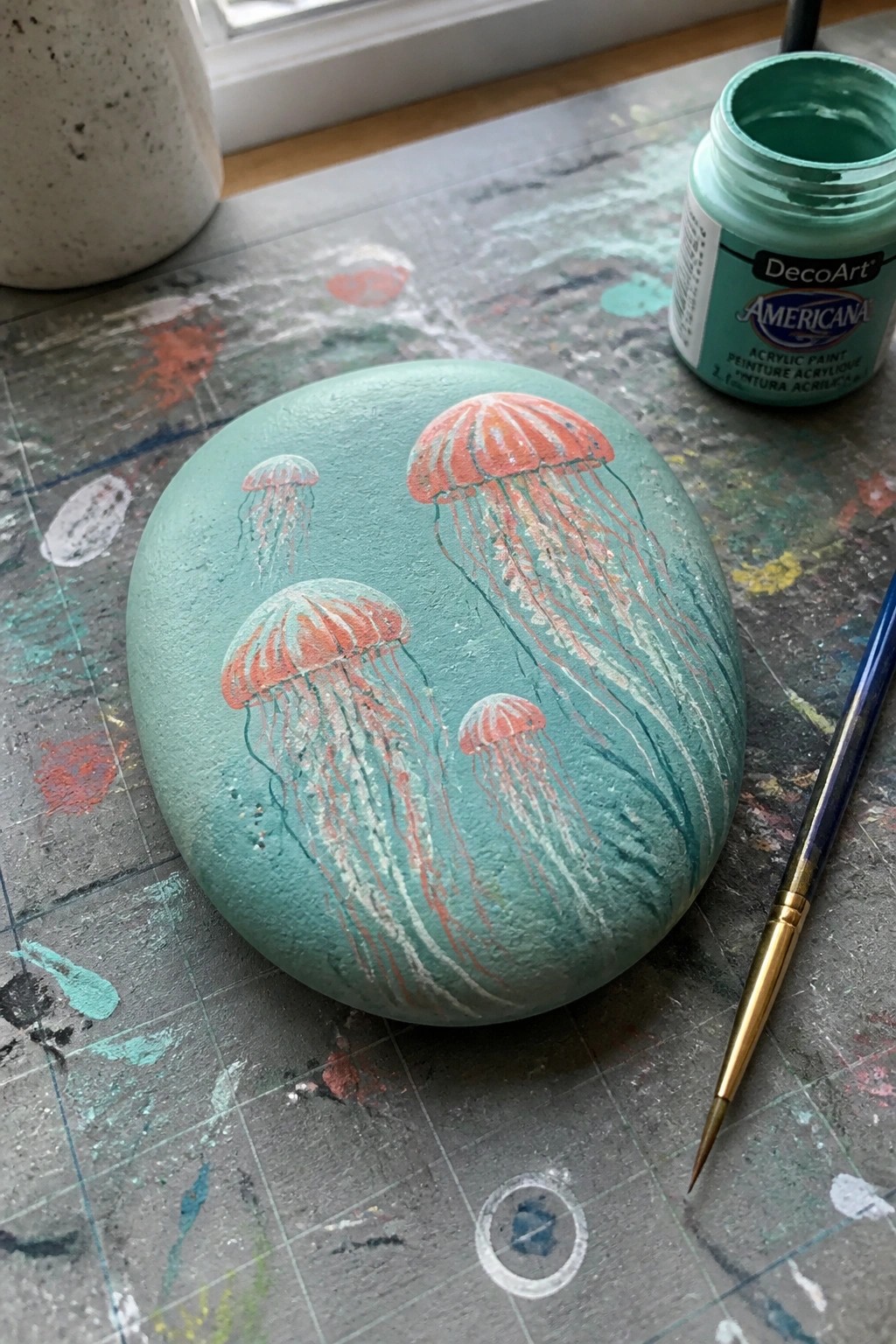 A smooth teal painted rock with several orange jellyfish designs rests on a paint-splattered surface next to a jar of green paint and a brush.