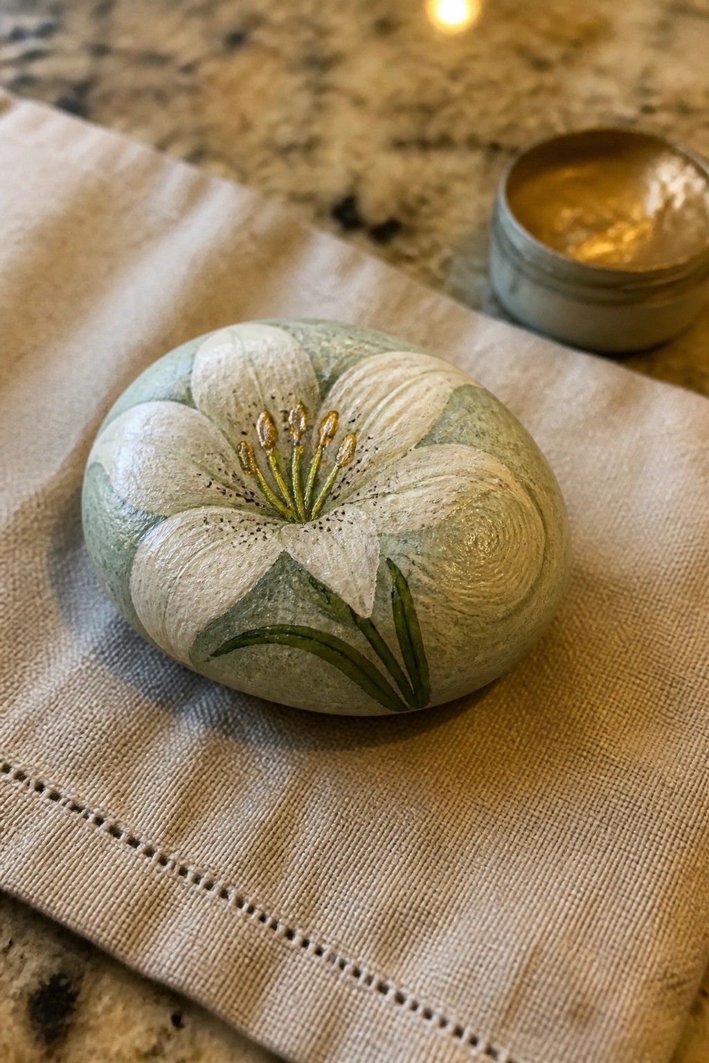 Painted green stone with white lily flower on beige fabric beside gold paint tin.
