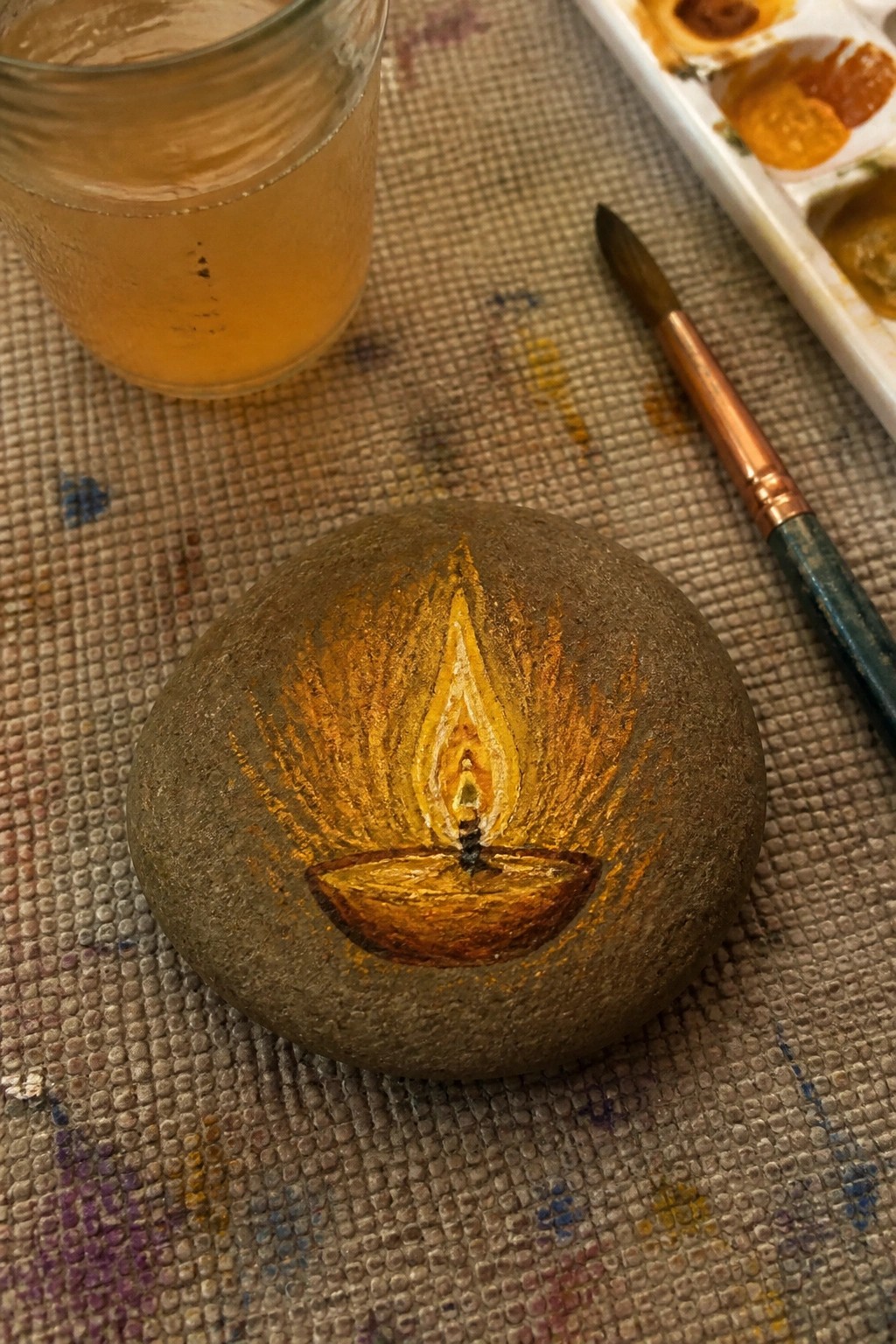 Painted stone with yellow diya flame, beside jar, brush, and palette on fabric.