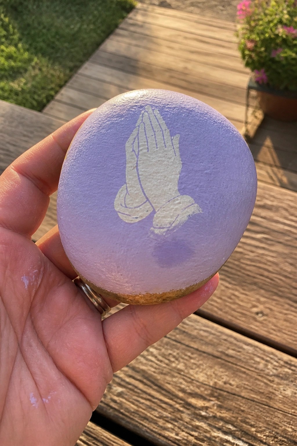 Hand holding light purple stone with white praying hands on wooden deck outdoors