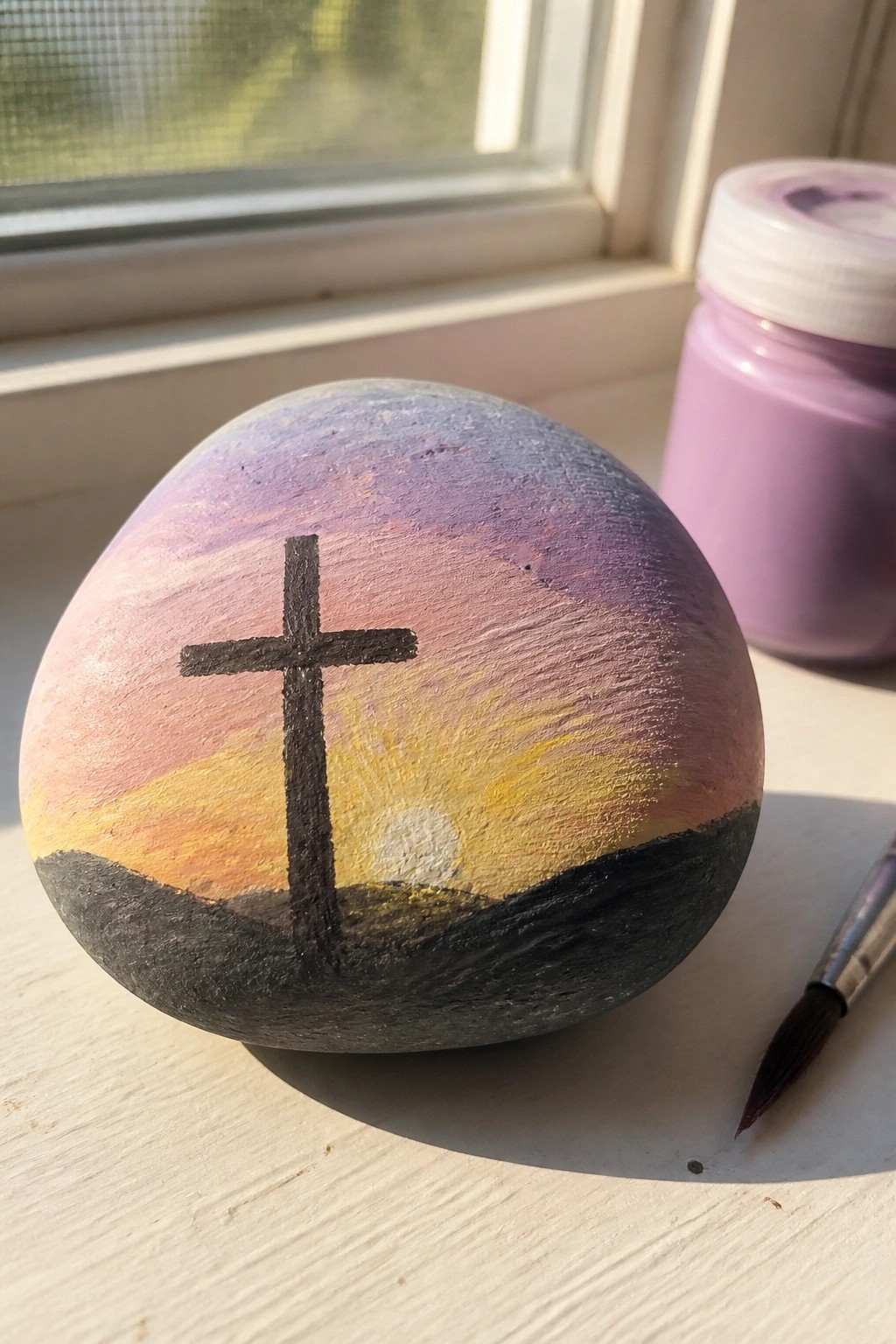 Painted rock with black cross over purple-pink sunset, paint jar and brush nearby