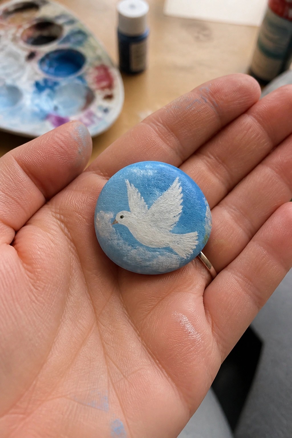 A hand holds a small round rock painted light blue with a white dove flying across clouds.