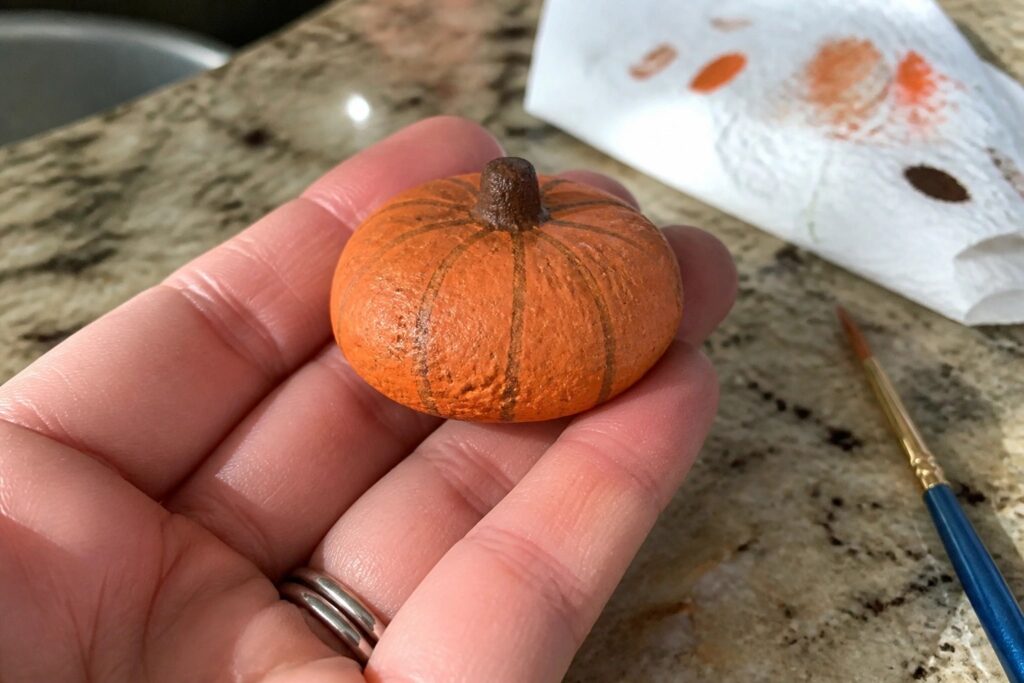 13 Cozy Fall Rock Painting Ideas Inspired by Pumpkins and Autumn Leaves Hand holding small orange painted pumpkin with brown stem near paintbrush on paper towel