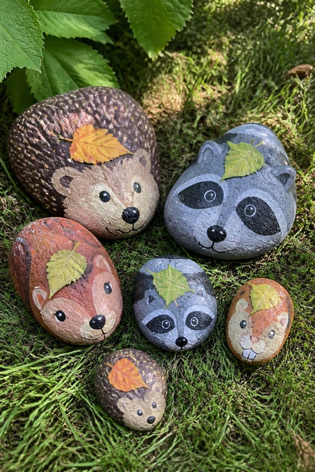 Six small painted rocks showing hedgehog, raccoon, and squirrel faces with autumn leaves placed on them, arranged on grass.