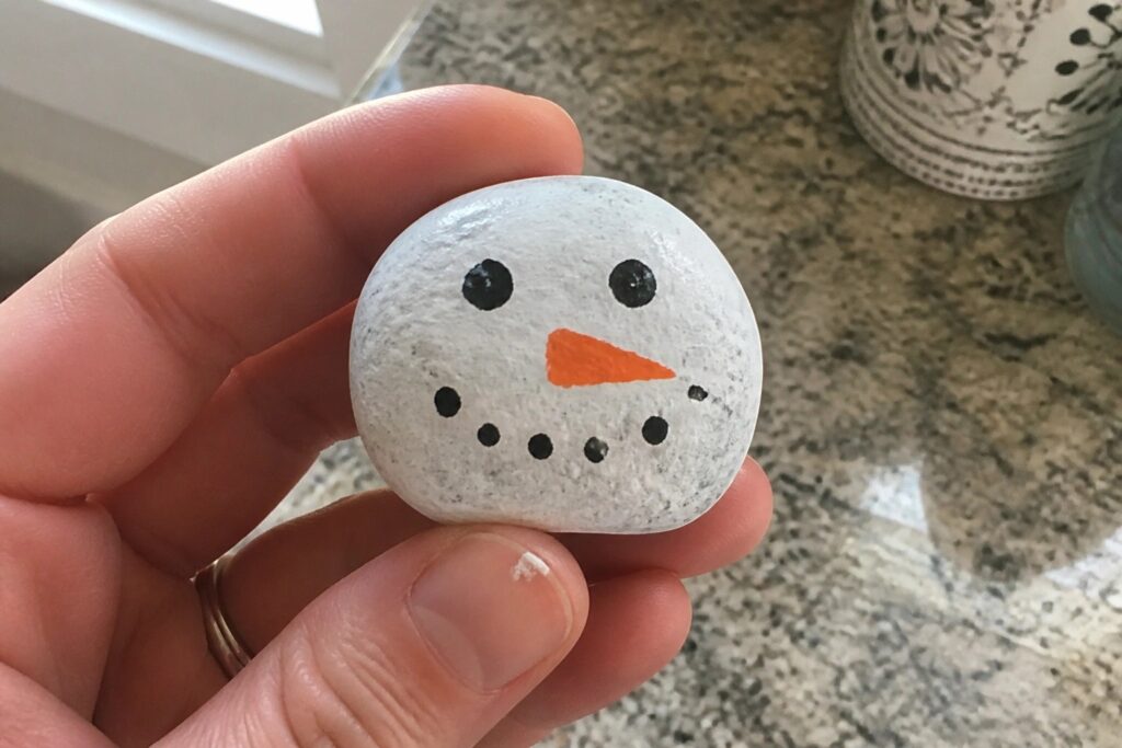 17 Festive Christmas Rock Painting Ideas for Simple Holiday Crafting Hand holding white painted rock snowman with black eyes and orange nose in kitchen