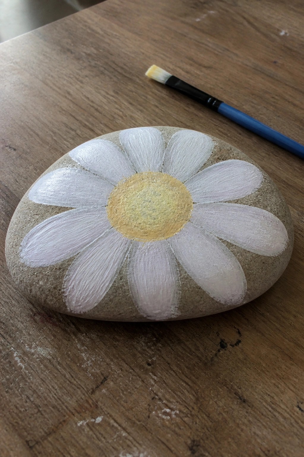 Painted stone daisy with white petals and yellow center beside paintbrush on wood