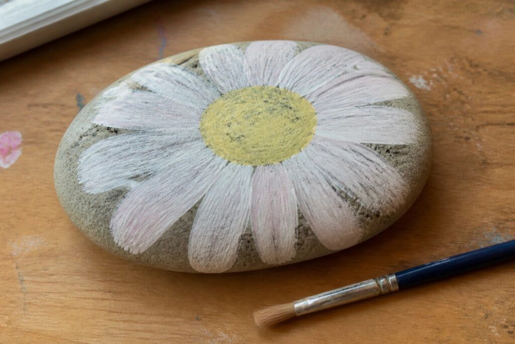 Painted stone daisy with white petals and yellow center beside paintbrush on wood
