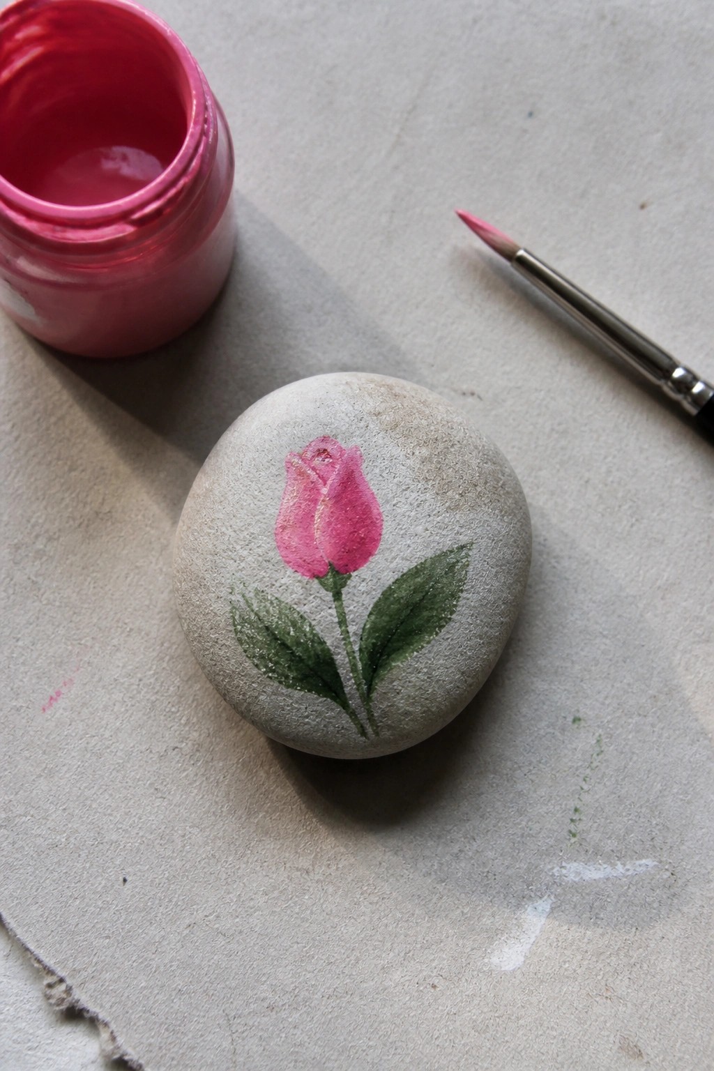 Gray stone painted with pink rose beside open pink paint jar and brush