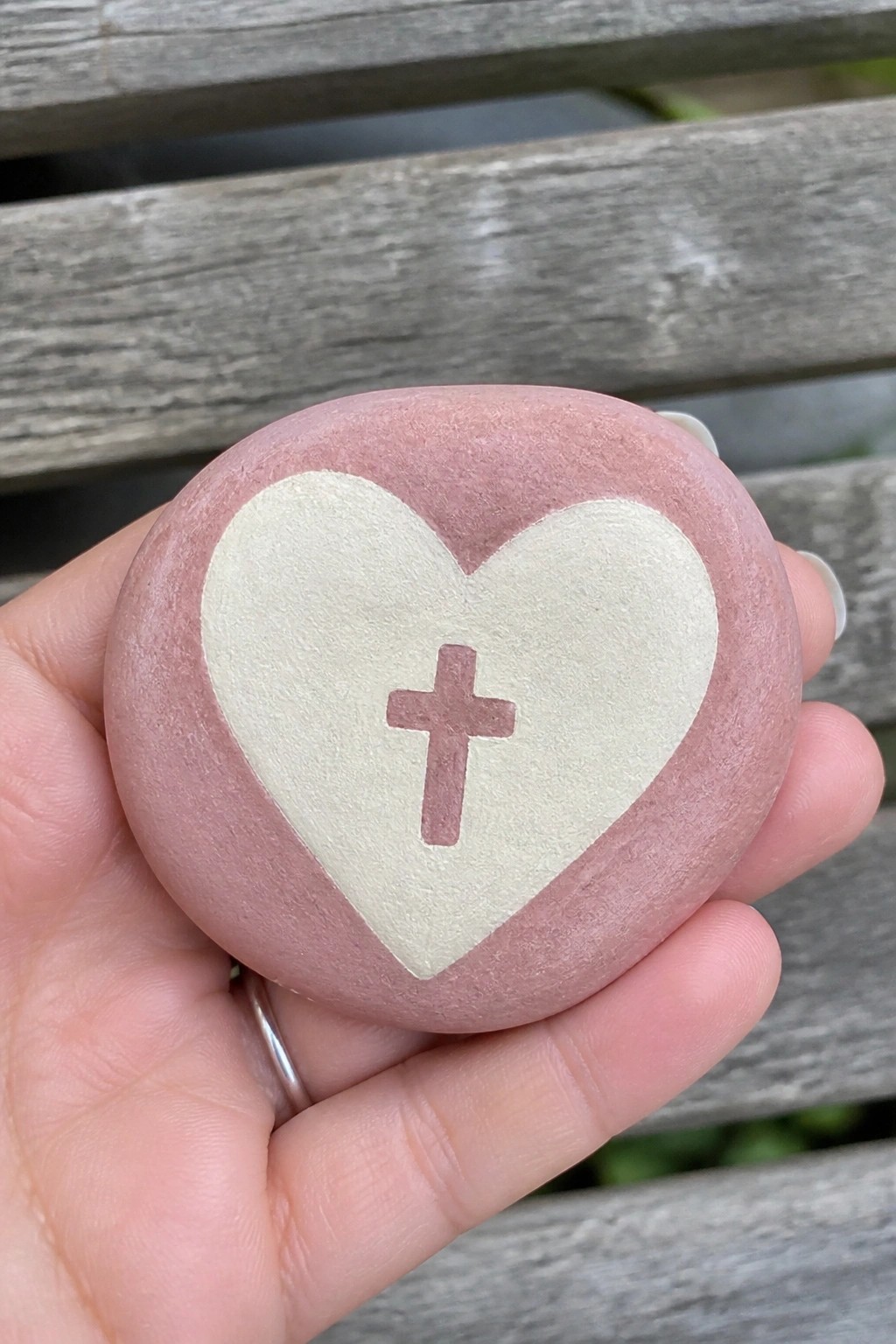 Hand holding pink stone with white heart and pink cross on wood bench