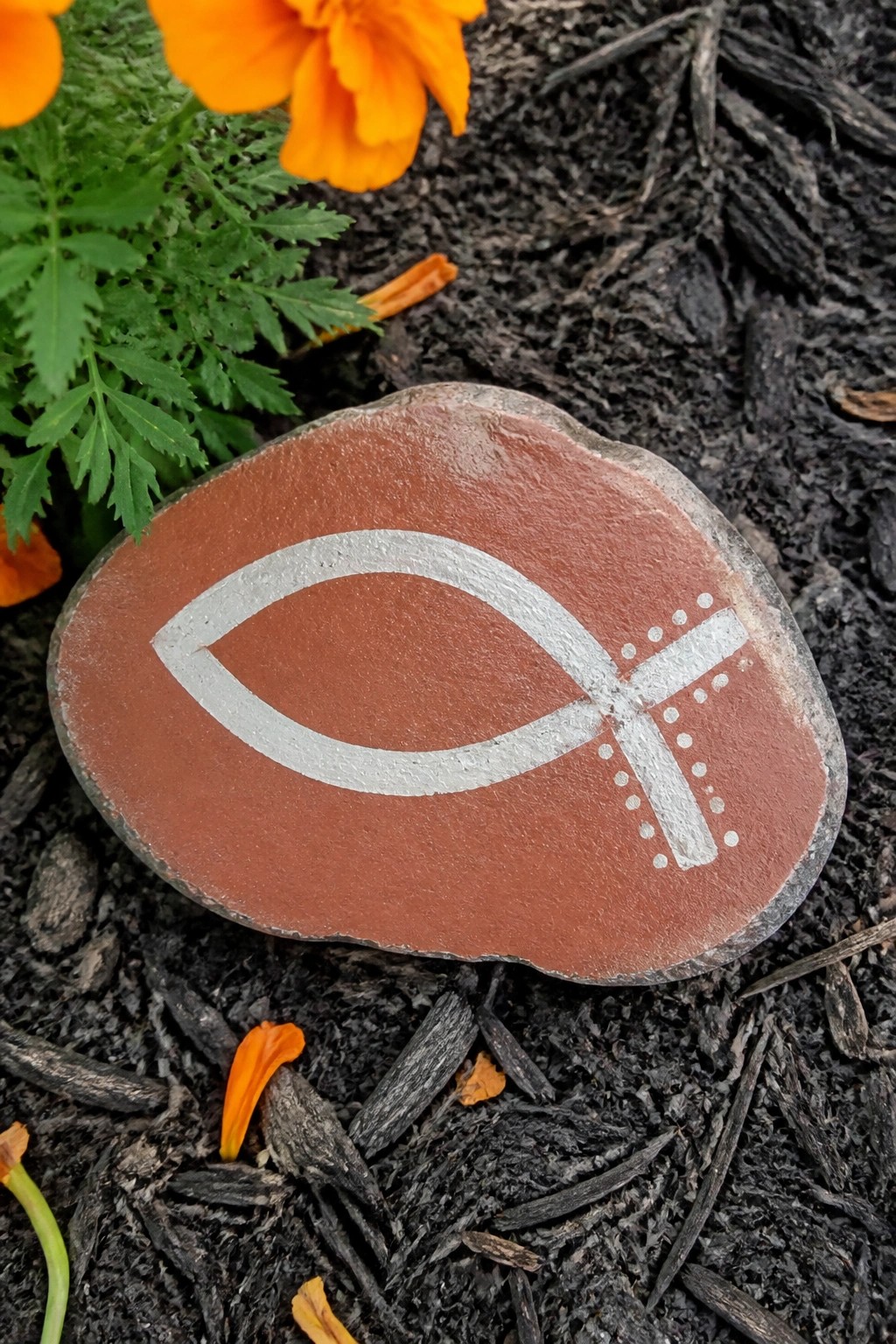 Reddish-brown rock with white fish and cross symbol on dark mulch near orange flower.