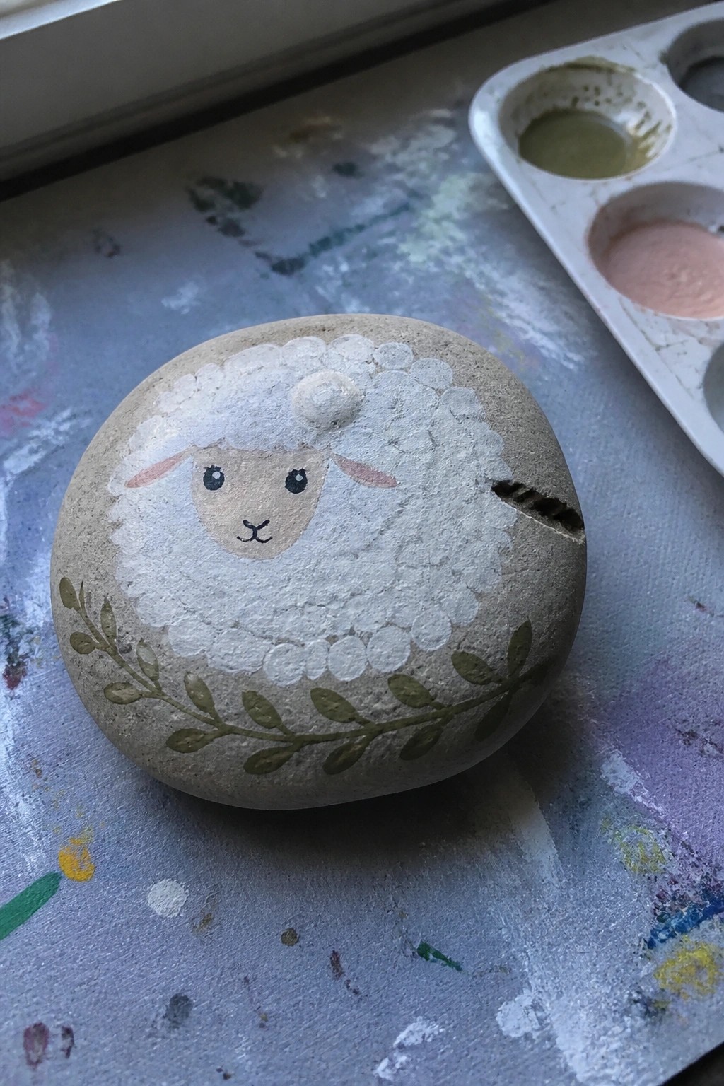 Painted sheep rock with white wool, face, pink ears, green leaves beside palette