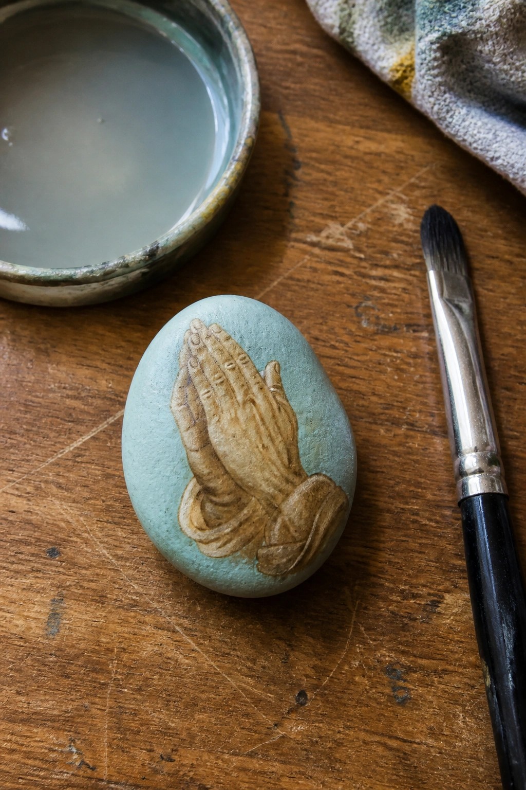 Painted turquoise stone with praying hands beside bowl and brush on wood