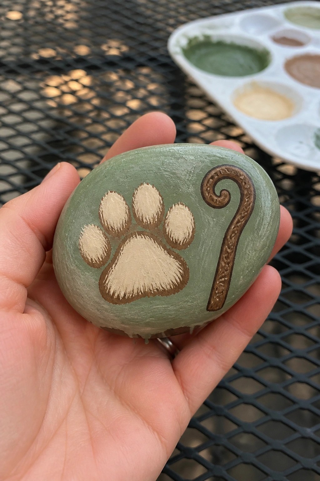 Hand holding light green rock painted with dog paw print and brown shepherd's staff