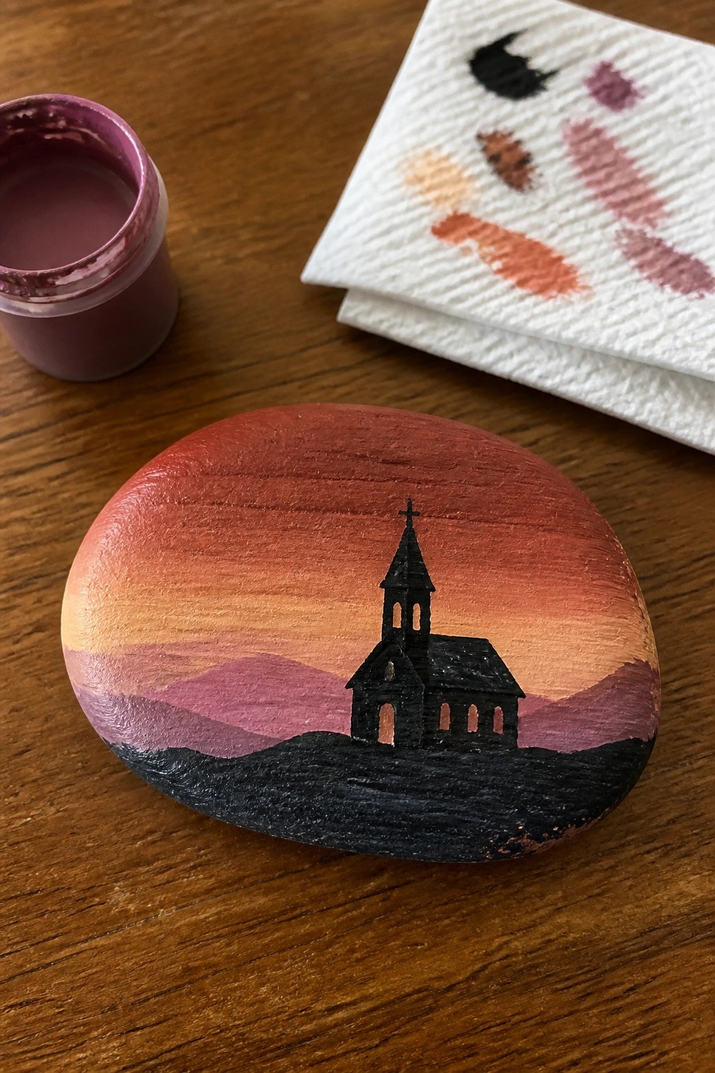 Painted rock with black church silhouette on orange-purple sunset mountains beside paint jar and towel.