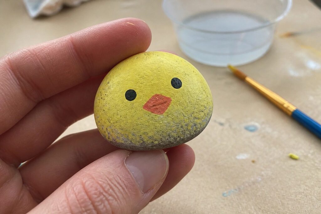 Yellow painted rock resembling a chick with black eyes and orange beak on wooden table.