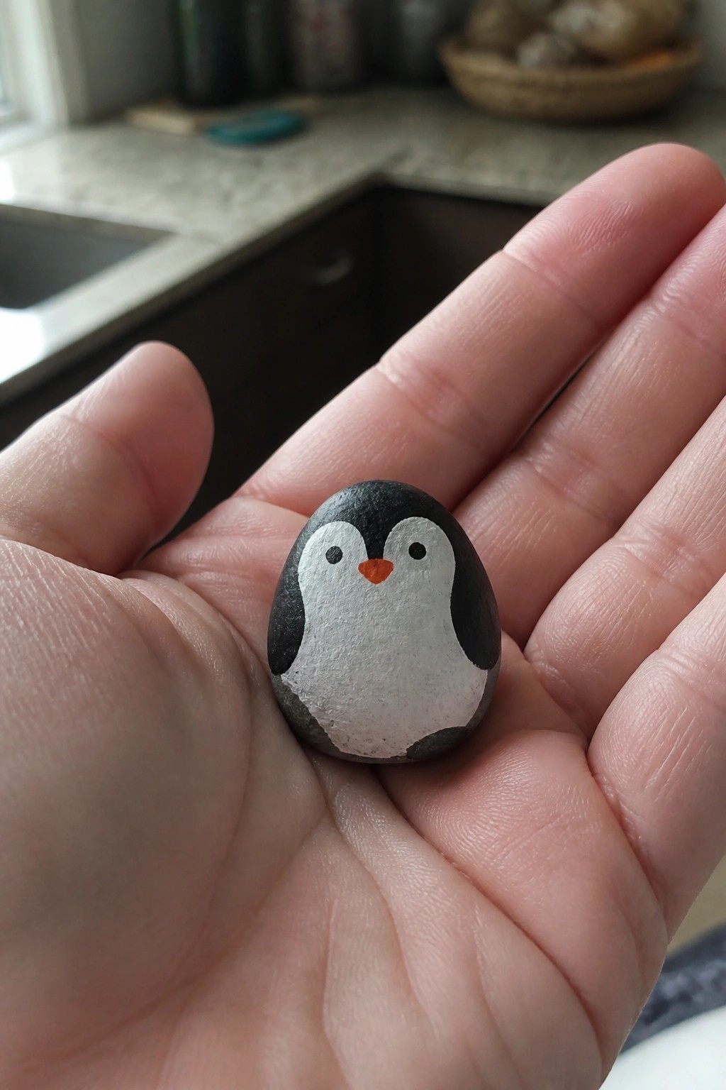 Open hand holding small oval rock painted as black-and-white penguin with orange beak.