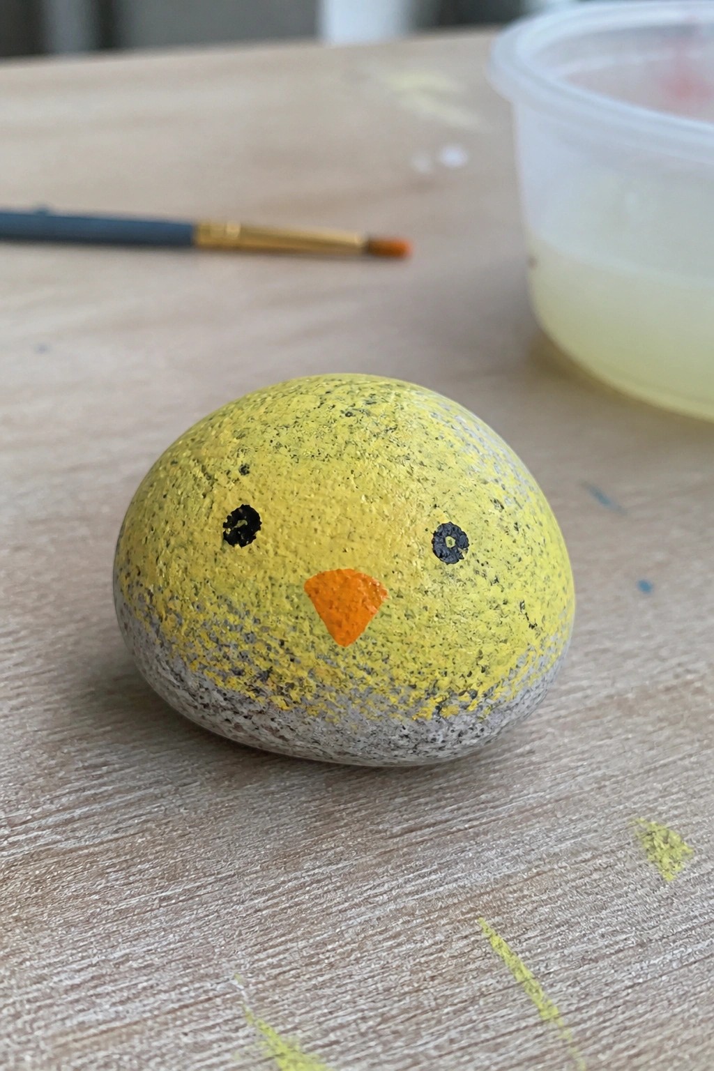 Yellow painted rock resembling a chick with black eyes and orange beak on wooden table.