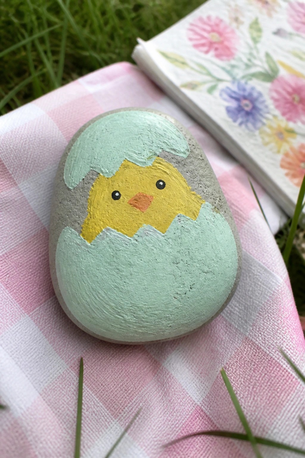 Painted rock of yellow chick hatching from cracked light green egg on pink checkered cloth