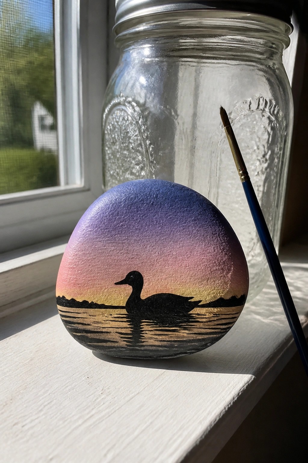 Painted rock with duck silhouette at sunset beside paintbrush and glass mason jar on sill