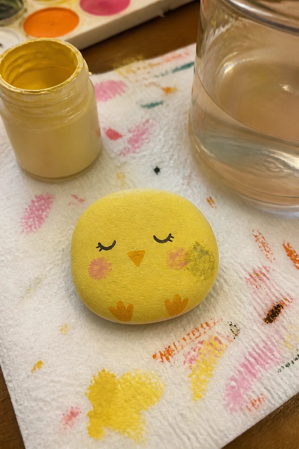 Yellow chick painted on rock on paint-stained paper towel beside yellow paint jar