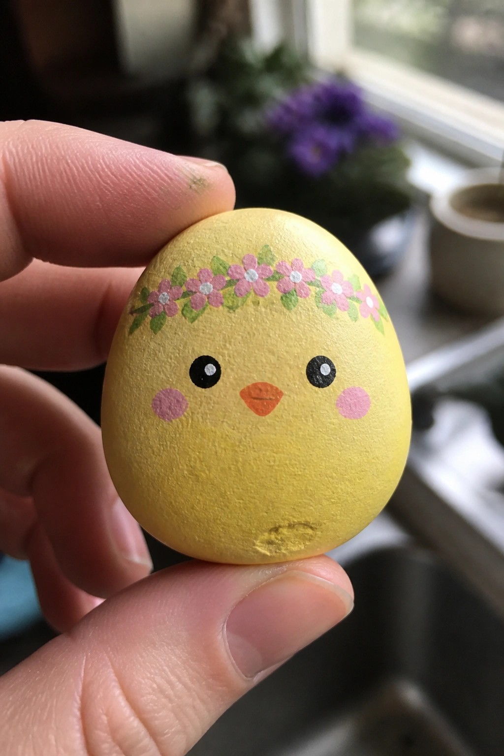 Hand holding yellow egg painted as chick with black eyes, orange beak and flower crown