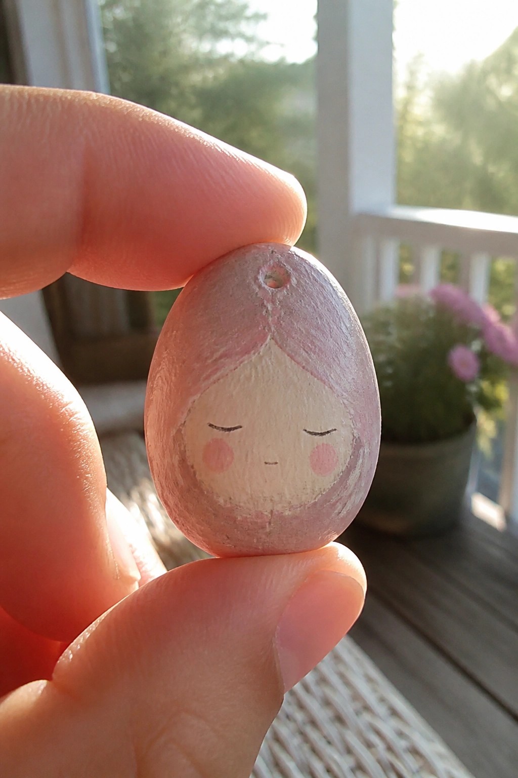 Fingers holding a small egg painted with pink hair, closed eyes, and cheeks.