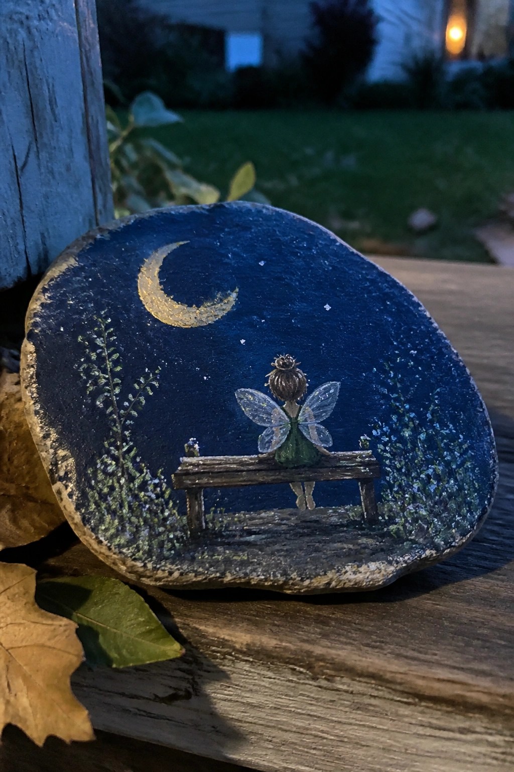 Painted rock with fairy in green dress on bench under golden crescent moon and stars.