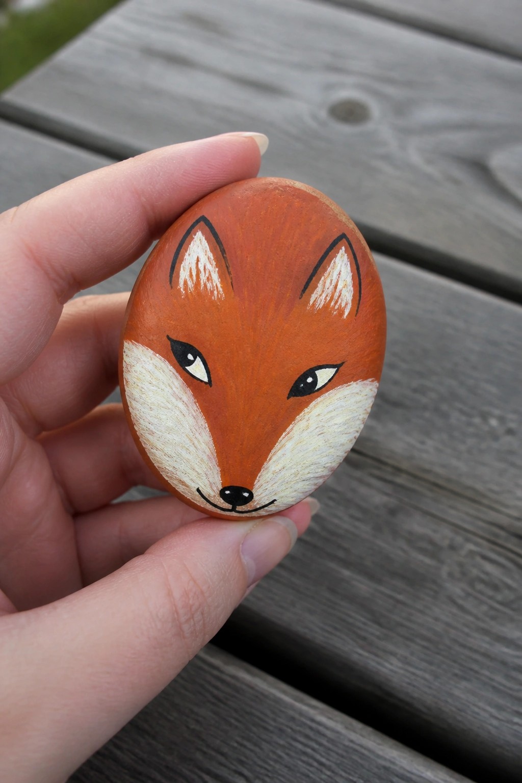 Hand holding oval rock painted as fox with orange fur and white muzzle