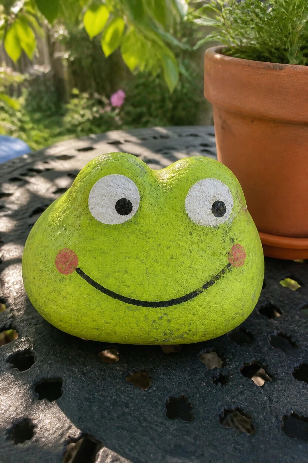 Green painted frog-faced rock with white eyes and smile on metal table near potted plant.