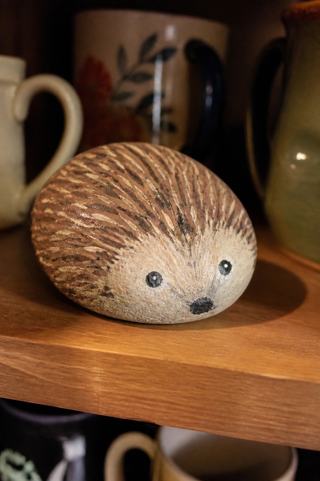 Painted hedgehog stone on wooden shelf among ceramic mugs and pitcher