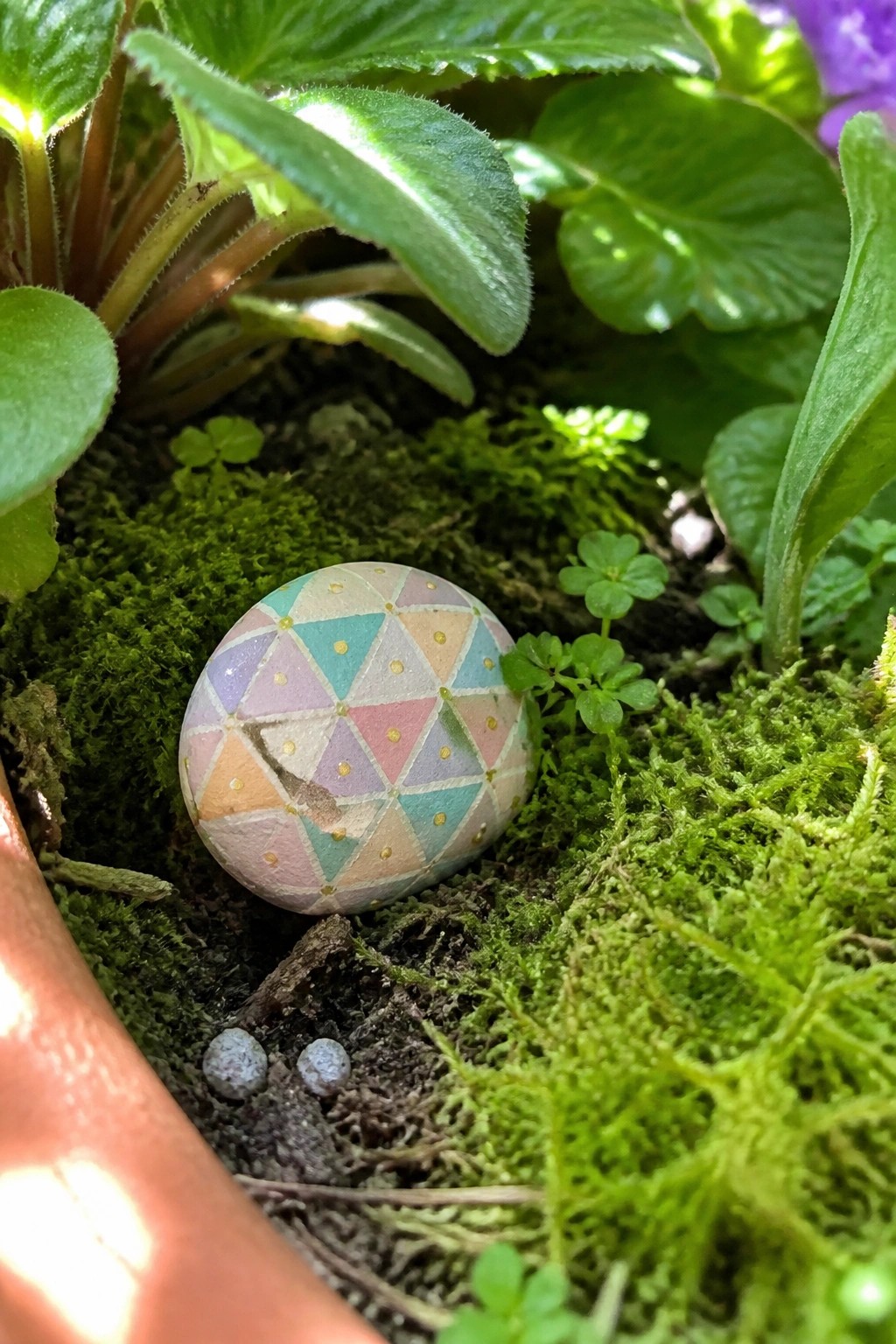 Painted stone with colorful geometric triangles and dots on green moss in a pot.