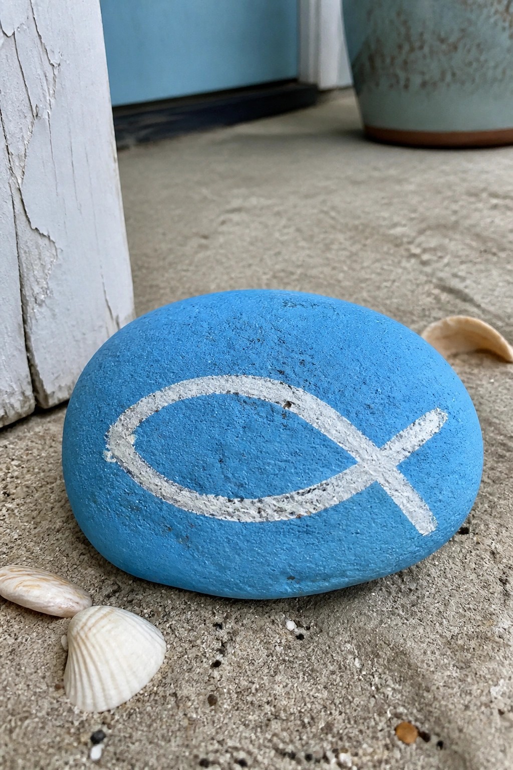 Blue painted rock with white fish symbol on sand beside seashells