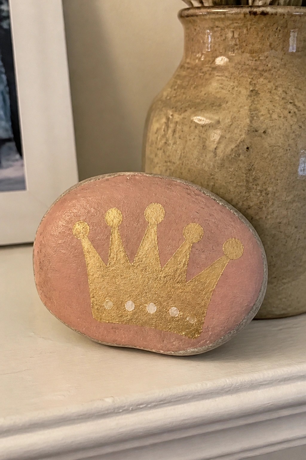 Peach painted rock with gold crown beside brown vase on white shelf
