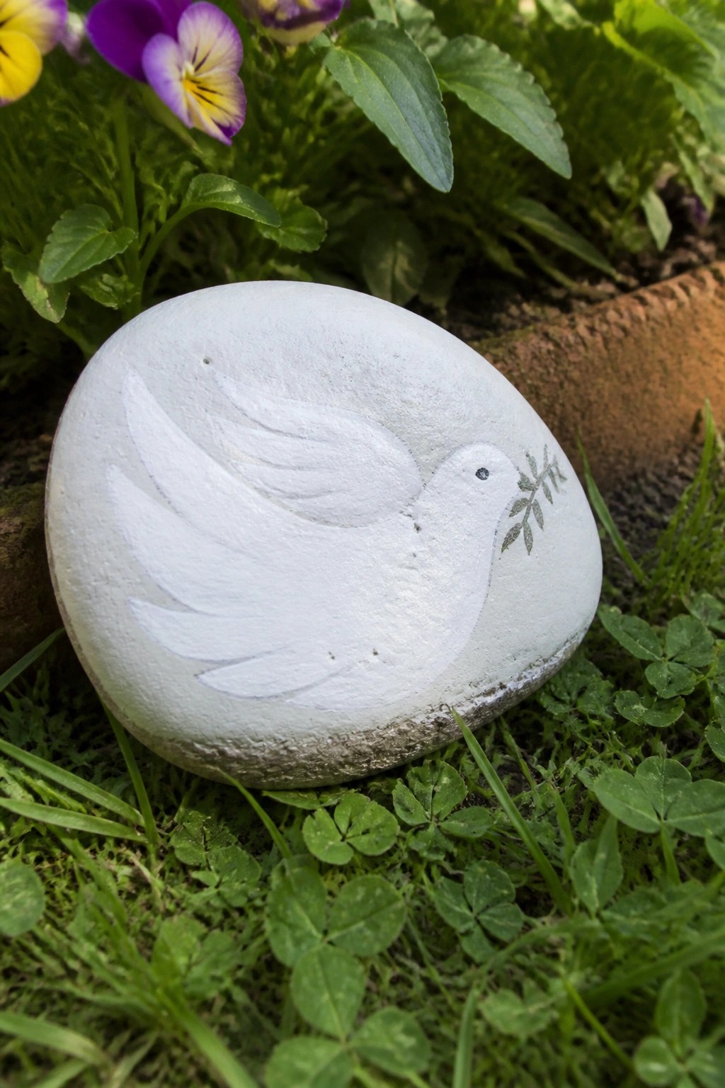 Painted white stone with dove and olive branch on grass amid flowers