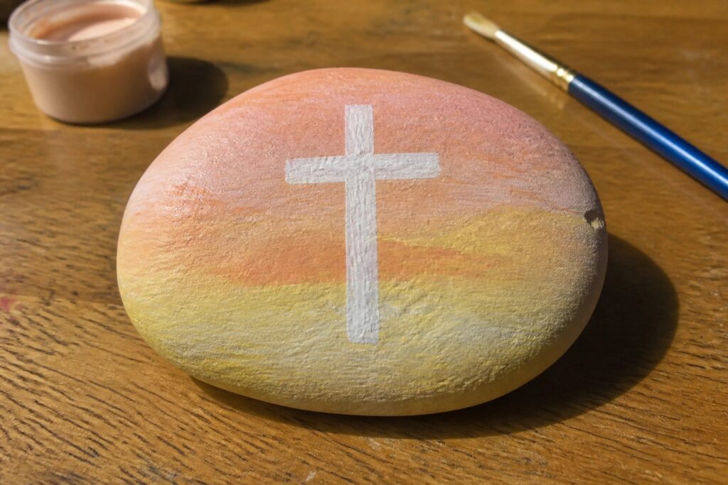 Painted rock with white cross on sunset gradient beside paint jar and brush