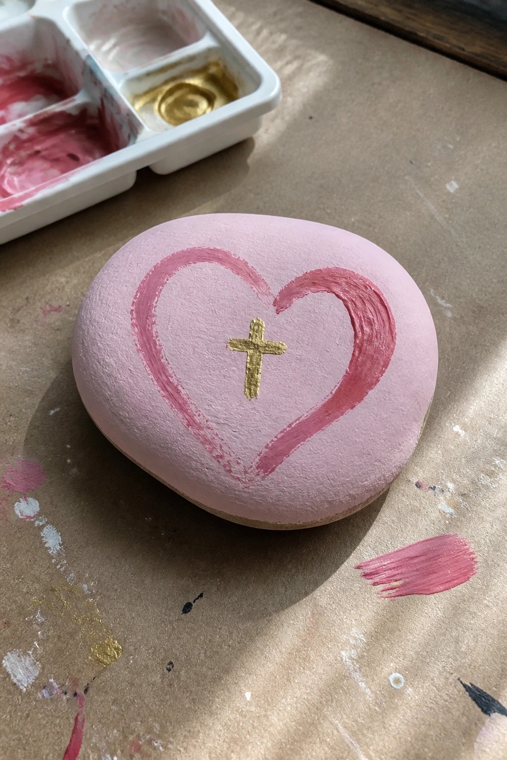 Pink rock with pink heart and gold cross beside paint palette