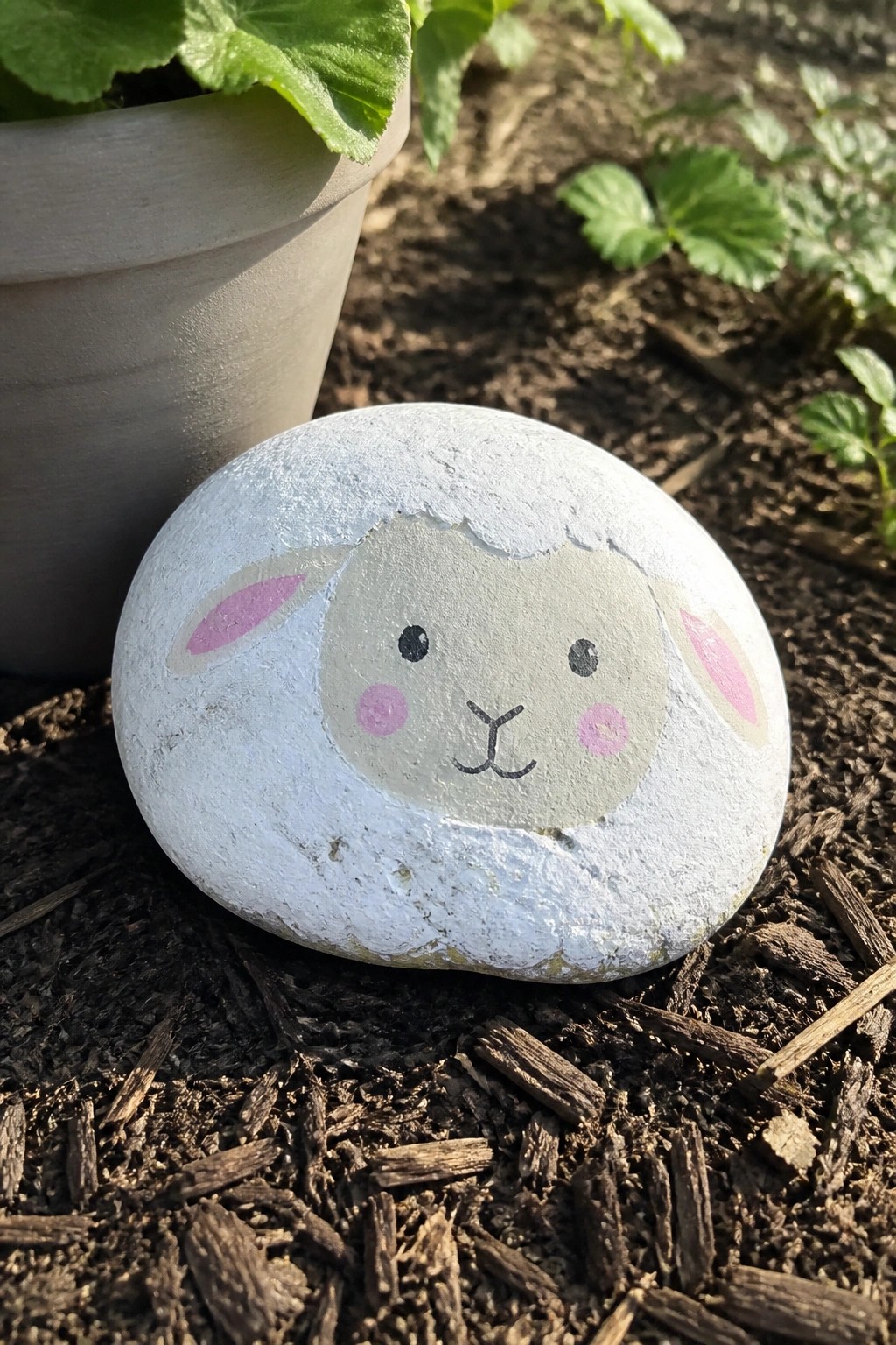 Painted white rock as sheep face with pink ears, cheeks on garden soil near plant