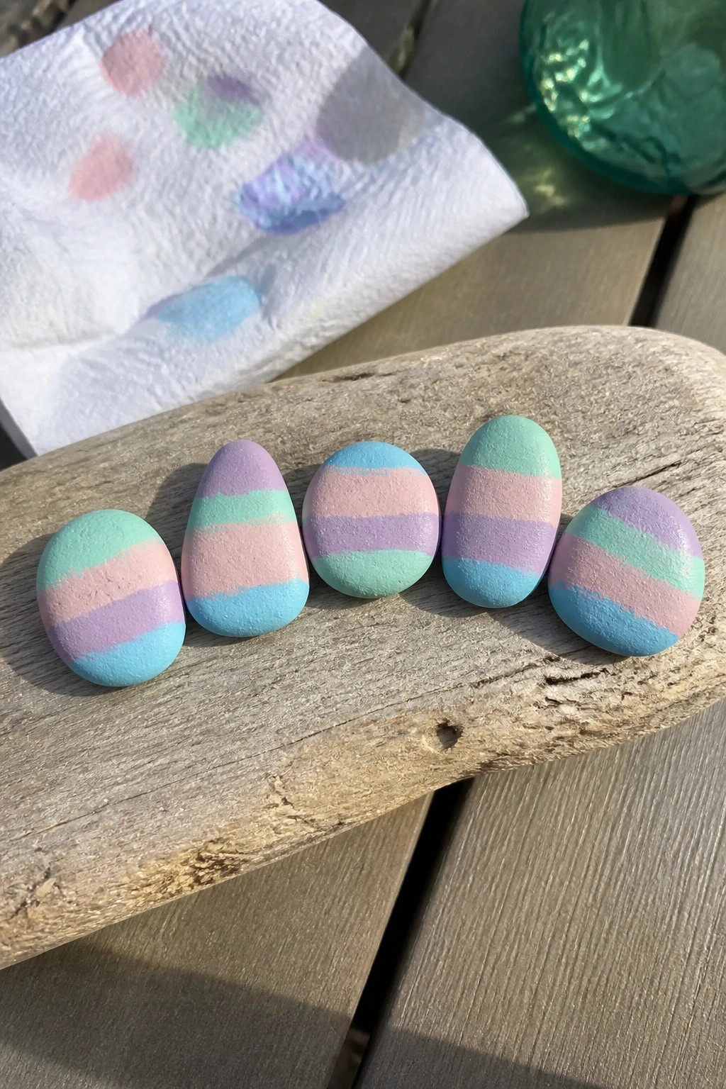 Five pastel-striped painted rocks lined up on weathered driftwood beside paint-smudged paper.