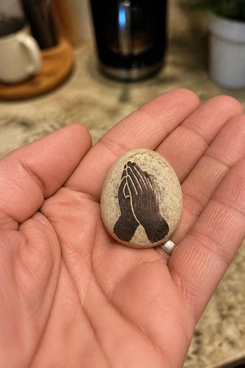Hand with silver ring holding oval stone painted with praying hands.
