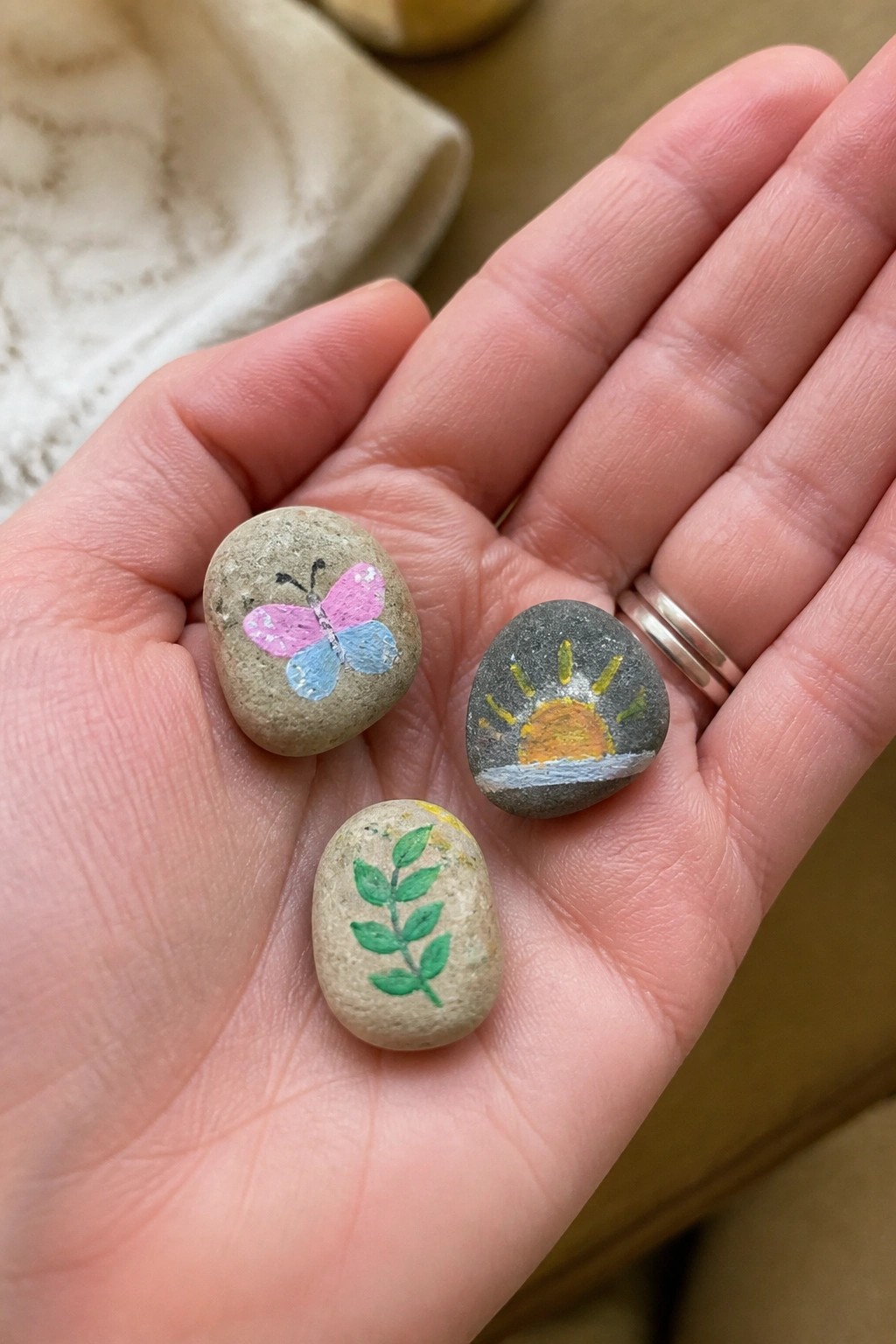Hand holding three painted rocks: pink butterfly, orange sun, green leaves