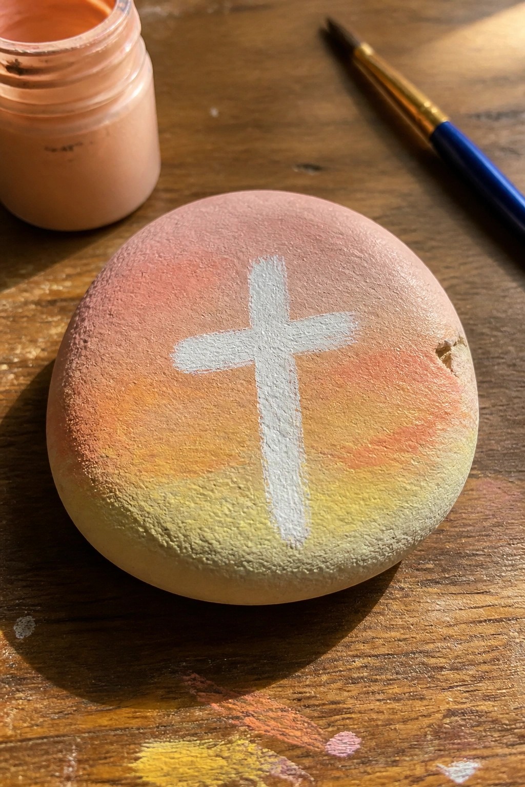 Painted rock with white cross on sunset gradient beside paint jar and brush