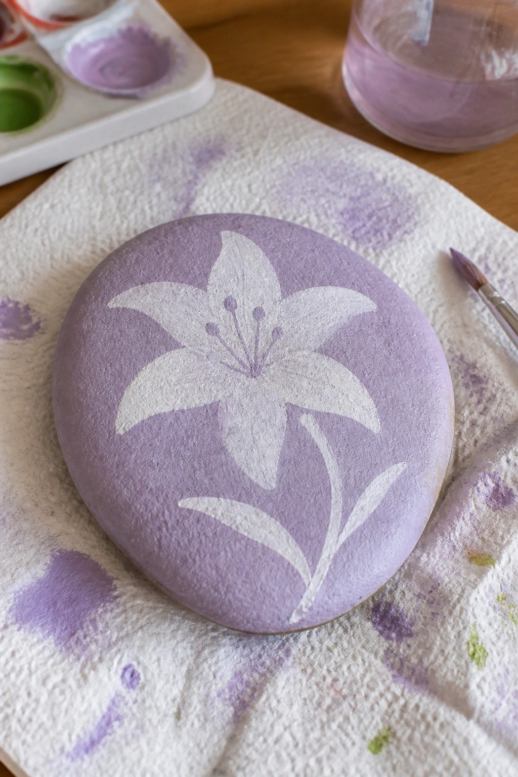 Painted purple rock with white lily on stained paper towel beside brush and palette.