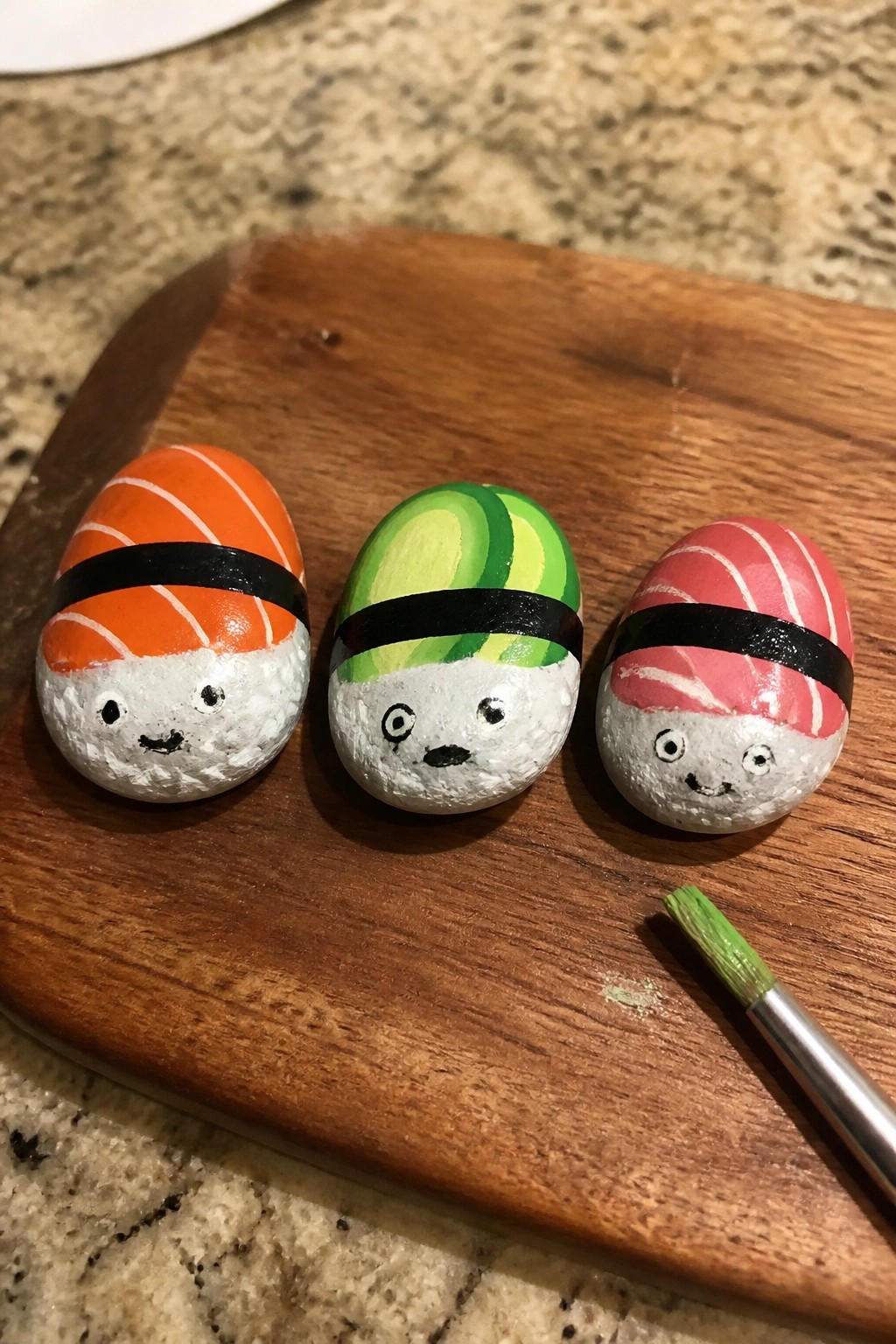 Three painted sushi rocks with faces on wooden board beside paintbrush