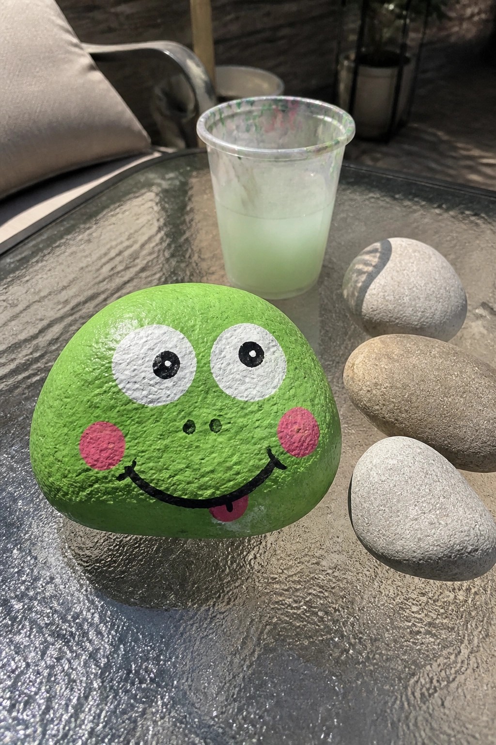 Green frog rock with pink cheeks and cartoon face on glass table near cup and stones