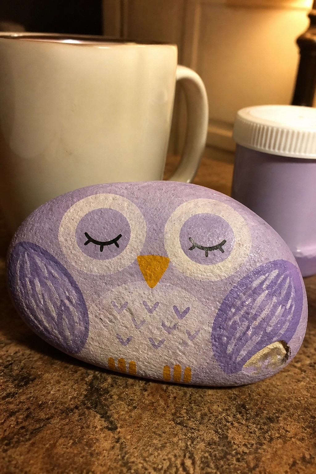 Purple painted owl rock with closed eyes, yellow beak, near white mug