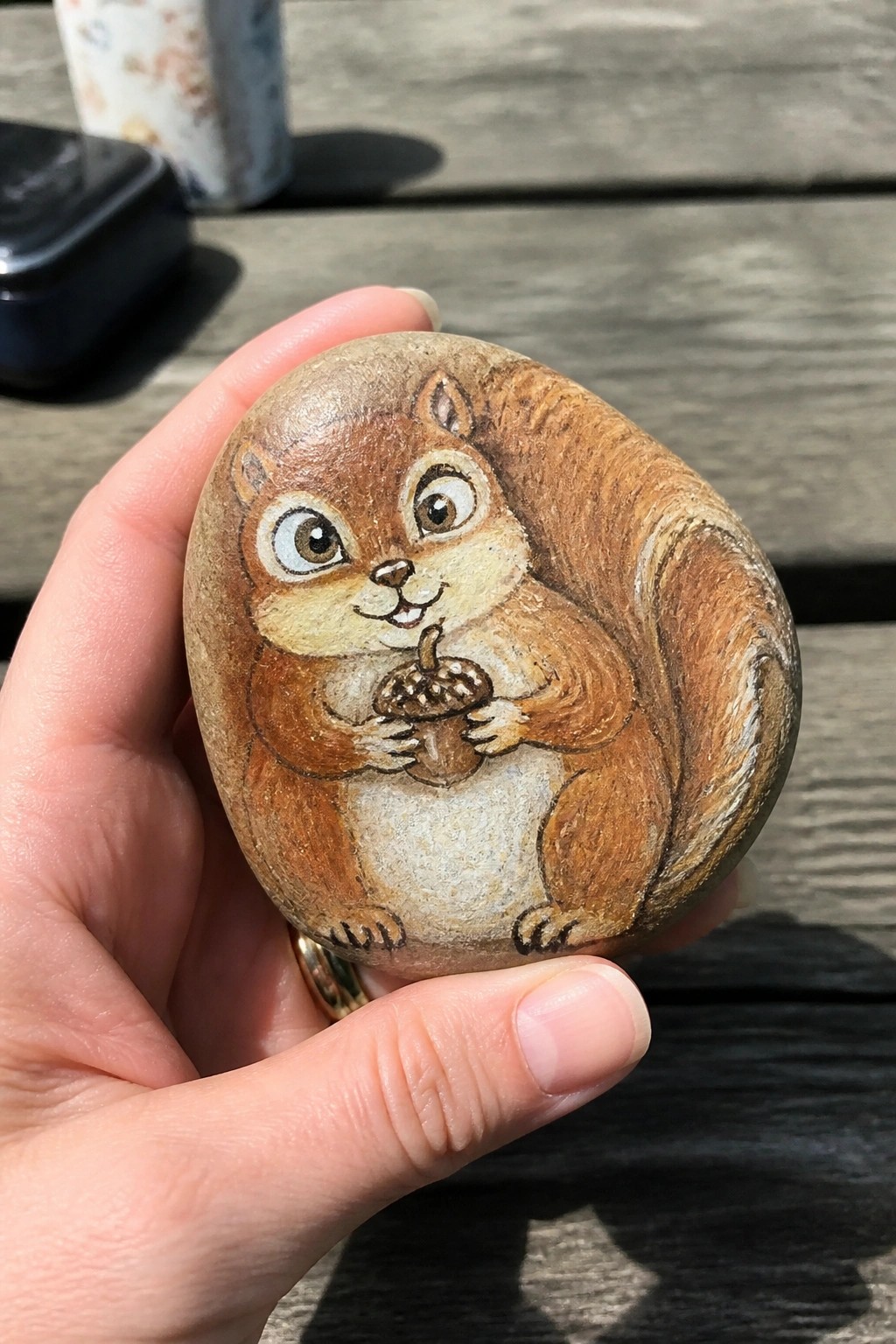 Hand holding painted rock of brown squirrel clutching acorn on wooden deck.