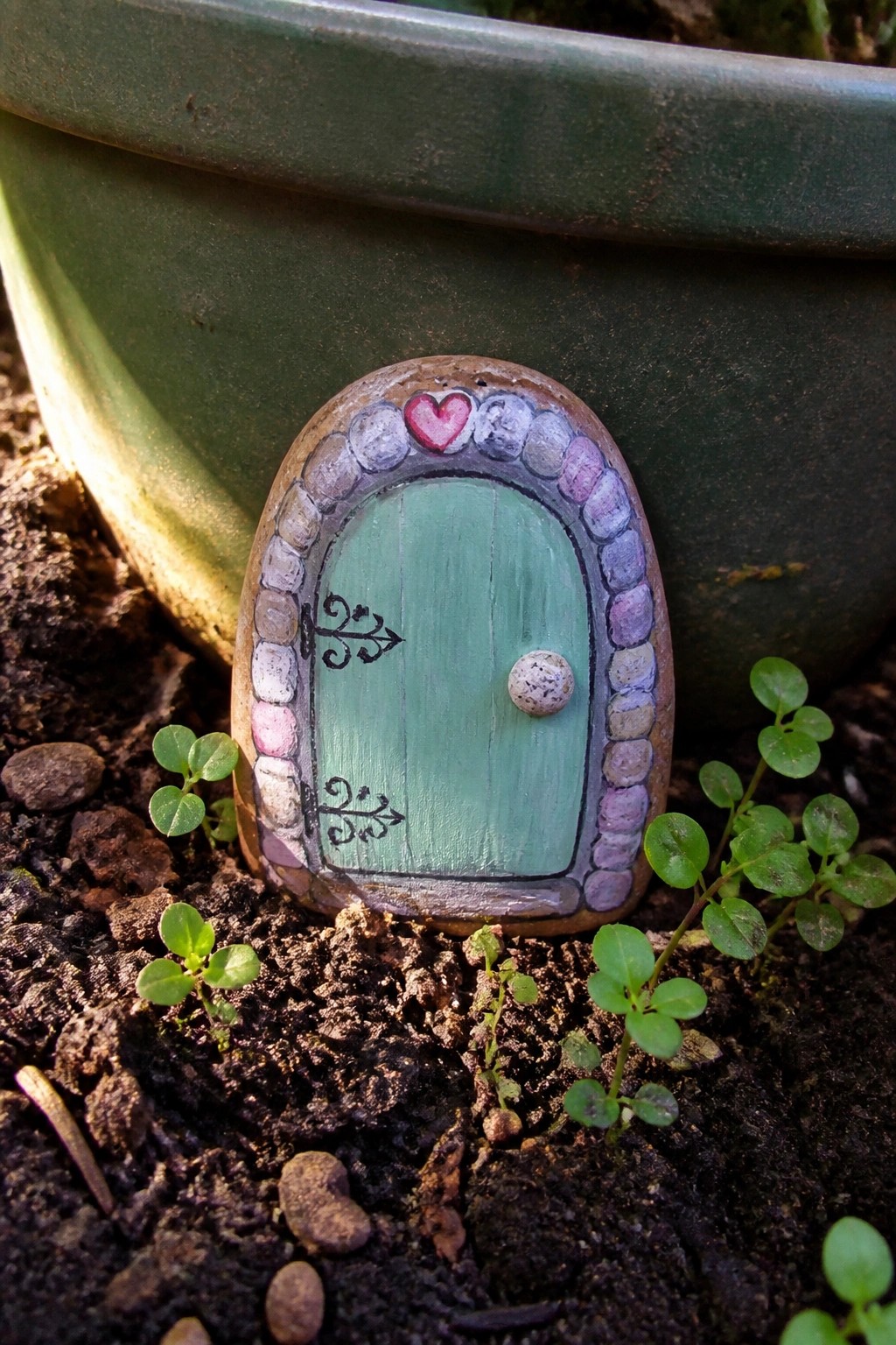 Painted rock fairy door with green panels, pink heart, in garden soil.