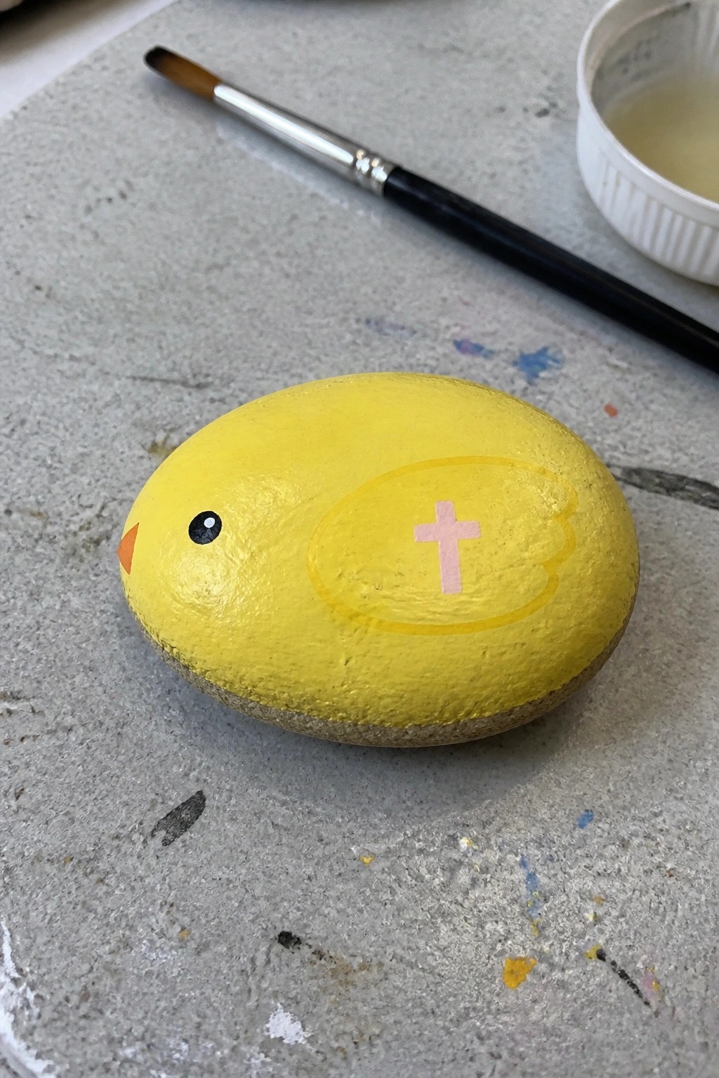 Yellow chick rock with pink cross wing beside paintbrush on gray surface